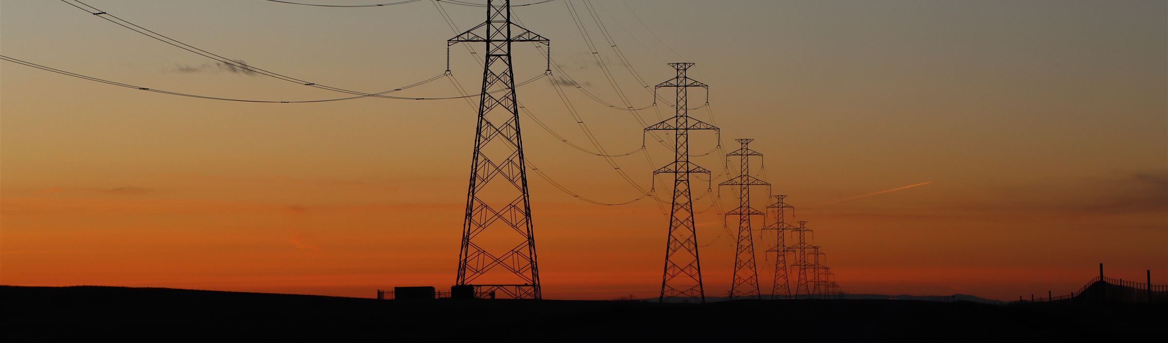 Row of lattice transmission towers silhouetted against an orange dusk sky